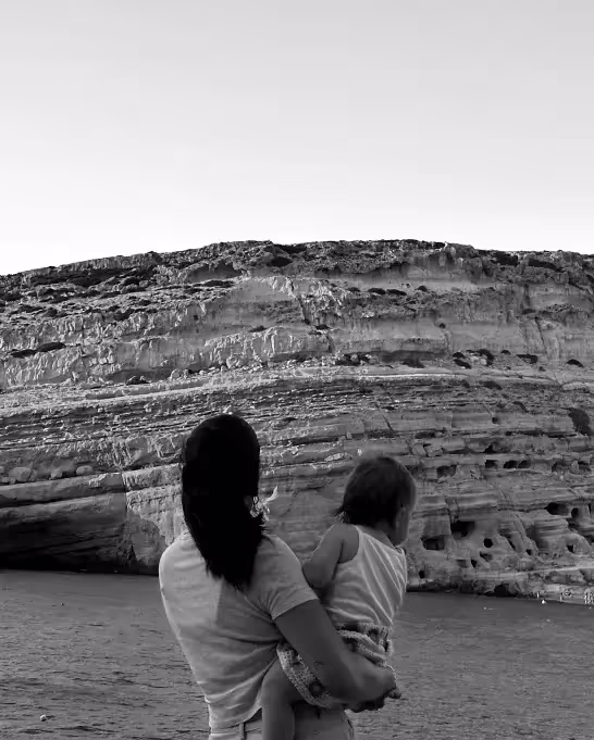 a woman holding a child on a beach near a cliff